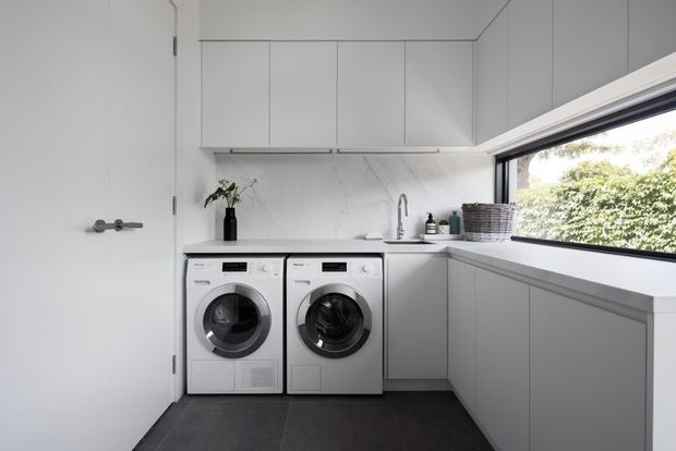 A bright, compact contemporary laundry room with two front-loading appliances beneath white cabinetry. The room features a white marble-look splashback, pale grey built-in cabinetry and benchtops, a stainless-steel tap and sink, dark tiled flooring, and a large horizontal window overlooking greenery.