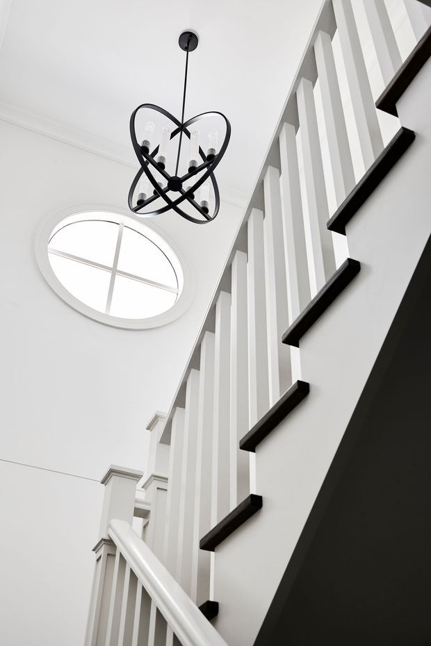 Upward-looking architectural detail of a bright white stairway with traditional balustrades and contrasting black stair treads. A large round window brings in natural light, while a sculptural black pendant chandelier hangs above the stair void. The space has crisp white trim, strong vertical lines, and a refined monochromatic palette.