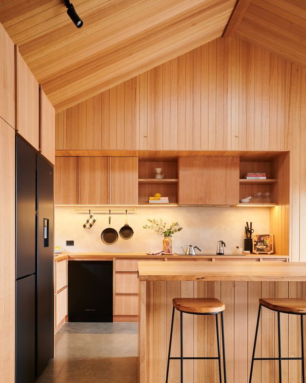 A compact, warm timber-lined kitchen with a pitched, fully panelled ceiling and walls. The kitchen features extensive vertical timber cabinetry and panelling, a pale stone or plaster-look splashback, integrated dark appliances, under-cabinet lighting, a timber island with matching panelled base, and two timber-topped black metal bar stools. Open shelving displays books and ceramics, while hanging cookware and a small vase add practical and decorative detail.