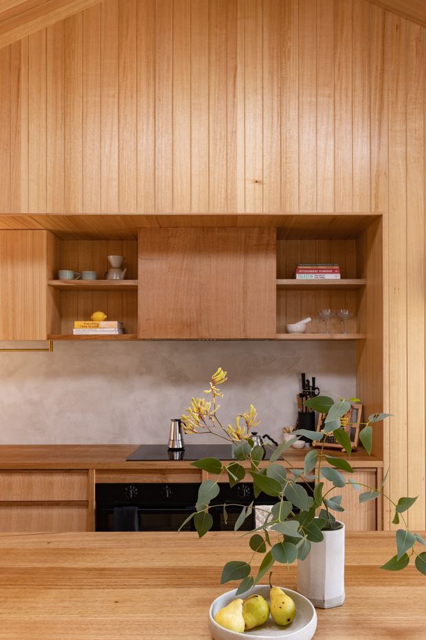 Warm, compact kitchen interior dominated by vertical timber wall panelling and matching built-in cabinetry. Open display shelving frames a central concealed rangehood or storage unit, with a muted concrete-look splashback, dark cooktop and oven, timber island or dining counter, and understated decorative objects and greenery in the foreground.