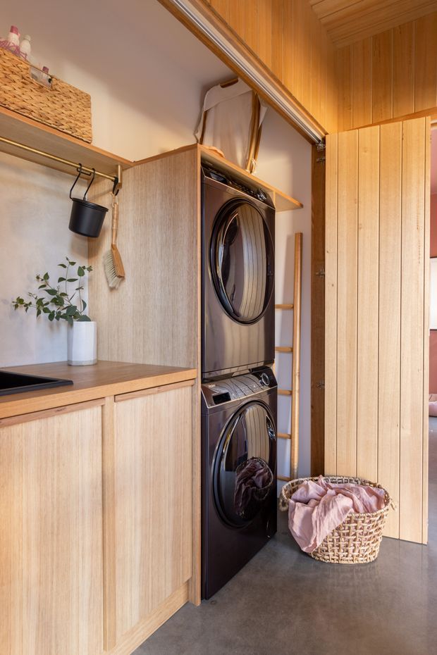 A compact, warmly finished laundry room with stacked black washer and dryer integrated beside light timber cabinetry. The room includes a timber benchtop with inset sink, open hanging storage, a woven laundry basket, ladder-style shelving, and vertical timber wall panelling. A doorway opens toward an adjacent room at the right.