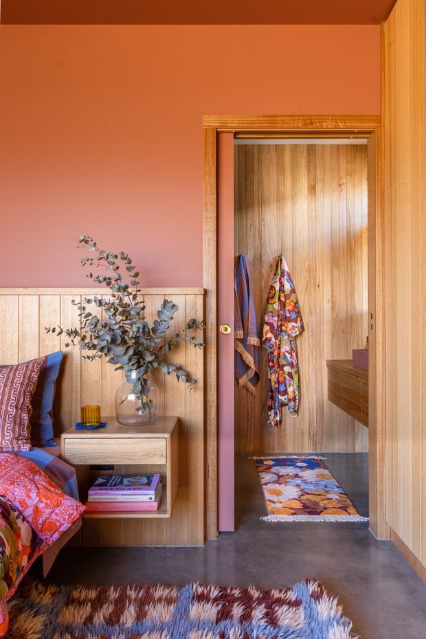 A colourful bedroom with a warm timber-panelled lower wall, terracotta-pink painted upper wall, built-in timber bedside table, patterned bedding, and layered multicoloured rugs. A timber-framed doorway opens to a compact timber-lined wardrobe or dressing area with hanging clothes and a low drawer unit.