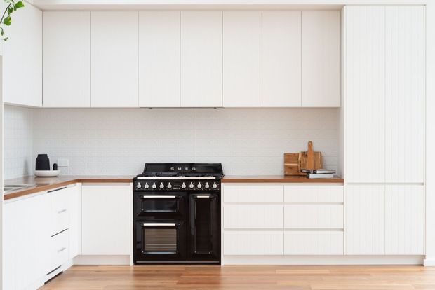 Bright, clean contemporary kitchen with white full-height cabinetry, a pale textured tile splashback, warm timber flooring, and a prominent black freestanding range cooker. The compact L-shaped layout includes continuous white benchtops, integrated storage, and simple vertical panel detailing.