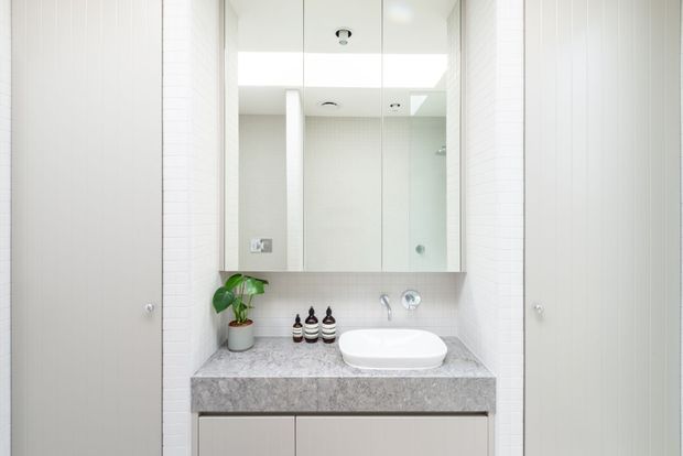 Bright, compact contemporary bathroom vanity with a pale stone benchtop, white vessel basin, mirrored wall cabinet, light grey cabinetry, and a glass-enclosed shower reflected in the mirror. White vertical wall tiling and integrated ceiling lighting create a clean, minimalist appearance.