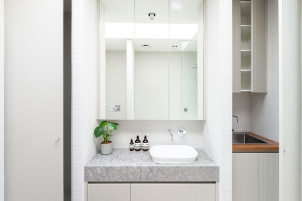 Bright, contemporary bathroom vanity with a wall-mounted mirror cabinet, white vessel basin, grey stone benchtop, pale cabinetry, white tiled walls, and integrated open shelving. A partially visible shower area and timber-toned ledge appear at the right.