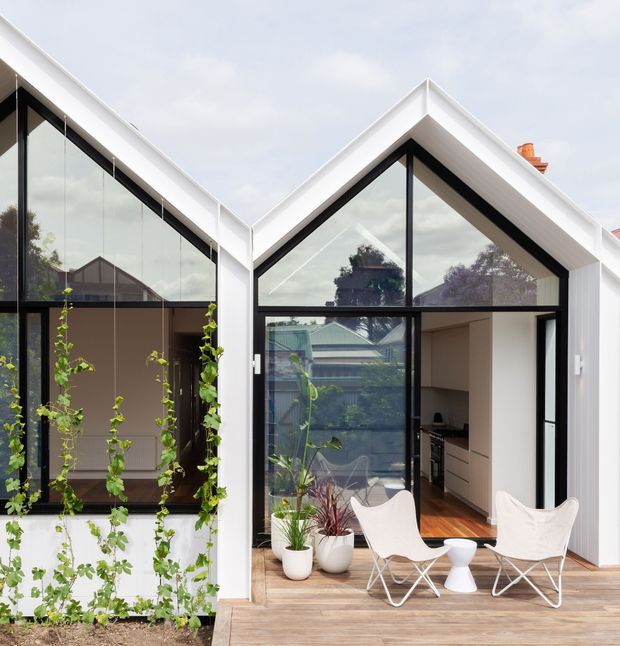 A contemporary white-clad residential extension with steep gabled forms, black-framed floor-to-ceiling glazing, and a timber deck. The open glazed living area includes two white sling chairs and a small side table, with potted plants and climbing greenery around the terrace. A compact kitchen is visible inside, while the adjoining glazed wing creates a bright indoor-outdoor connection.