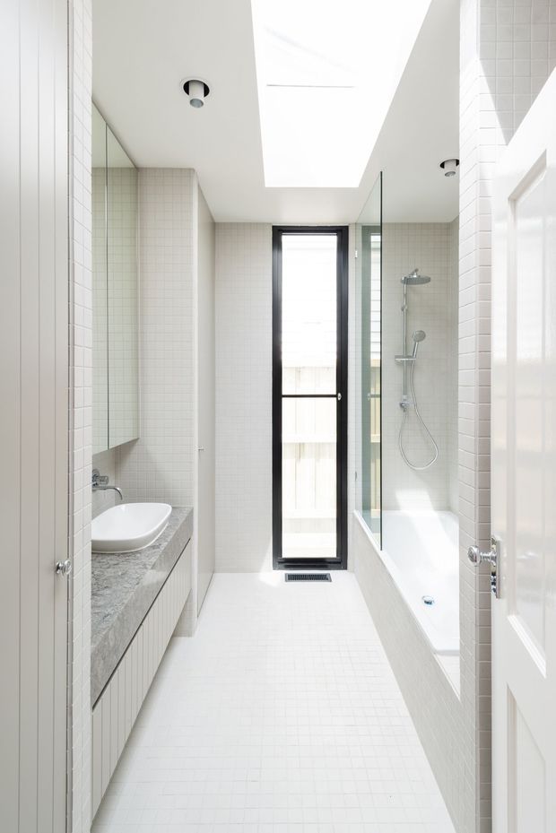 Bright, narrow contemporary bathroom with white small-format tiled walls and floor, a freestanding-style bathtub with clear glass shower screen, wall-mounted shower fittings, a grey stone vanity benchtop with white vessel basin, black-framed vertical window, recessed ceiling lights, and a large skylight.