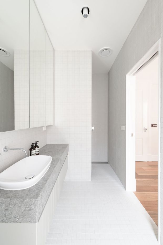 Bright, minimalist bathroom with a white vessel basin set into a grey stone benchtop, large mirrored wall cabinetry, white small-format tiled walls and floor, and a frameless shower area behind a tiled partition. A doorway opens to a timber-floored adjacent room.
