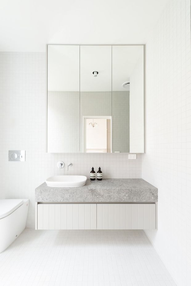 Bright minimalist bathroom featuring white small-format tiled walls and floor, a floating vanity with vertically fluted white cabinetry, a grey stone benchtop, white vessel basin, wall-mounted tapware, and a large three-panel mirrored cabinet. A freestanding white bathtub is partially visible at left.