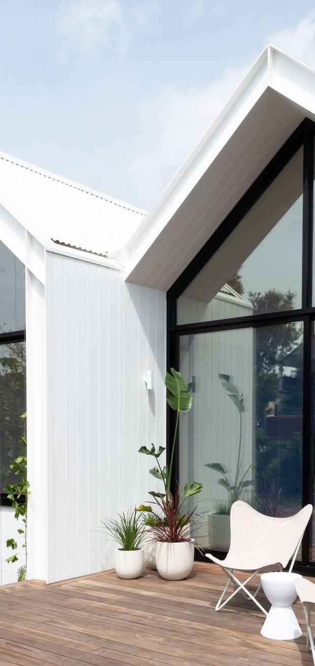 Exterior architectural photograph of a bright white gabled dwelling with vertical white cladding, a steep pitched roof, black-framed floor-to-ceiling glazing, and a timber deck furnished with a white sling chair and potted plants. The composition highlights the crisp gable junction and indoor-outdoor connection.