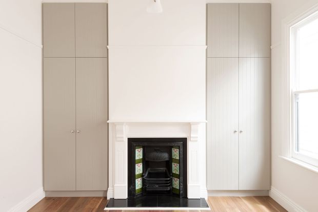 Bright, symmetrical room featuring a traditional fireplace with decorative green-and-white tilework, white mantel and chimney breast, full-height built-in cabinetry on both sides, pale walls, timber flooring, and a large sash-style window.