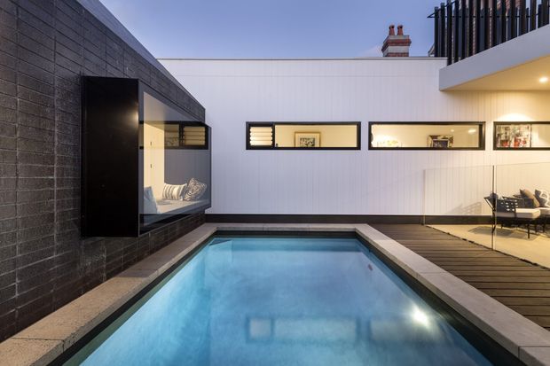 Contemporary urban courtyard with a narrow rectangular swimming pool surrounded by dark timber decking and pale stone coping. The space is enclosed by dark textured masonry and white vertical cladding, with integrated horizontal windows, a large glazed opening revealing an interior seating area, and an upper-level balcony with black vertical balustrading. Warm recessed lighting adds contrast to the cool blue pool water.