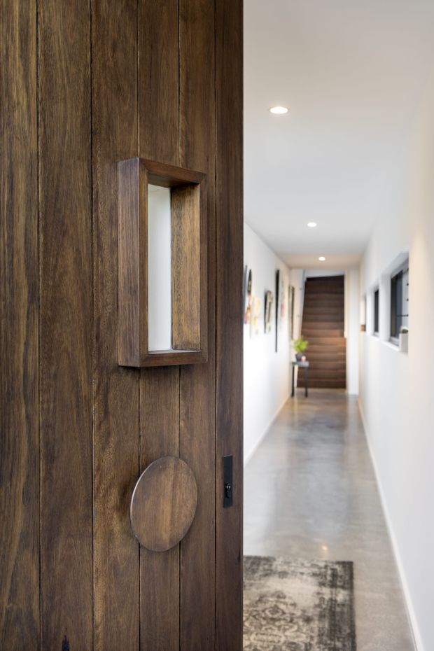 View through a distinctive dark timber pivot door into a long, bright hallway. The door has vertical timber panelling, a small inset glazed opening, and a circular timber detail. The hallway features white walls, recessed ceiling lights, polished grey concrete flooring, framed artwork, narrow windows, and a timber-clad stair at the far end.