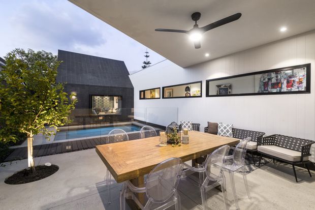 Covered outdoor dining and entertaining area beside a narrow swimming pool, with a large timber dining table, transparent dining chairs, patterned lounge seating, ceiling fan, recessed lighting, and framed wall art. The space opens toward a dark-clad structure and landscaped courtyard.