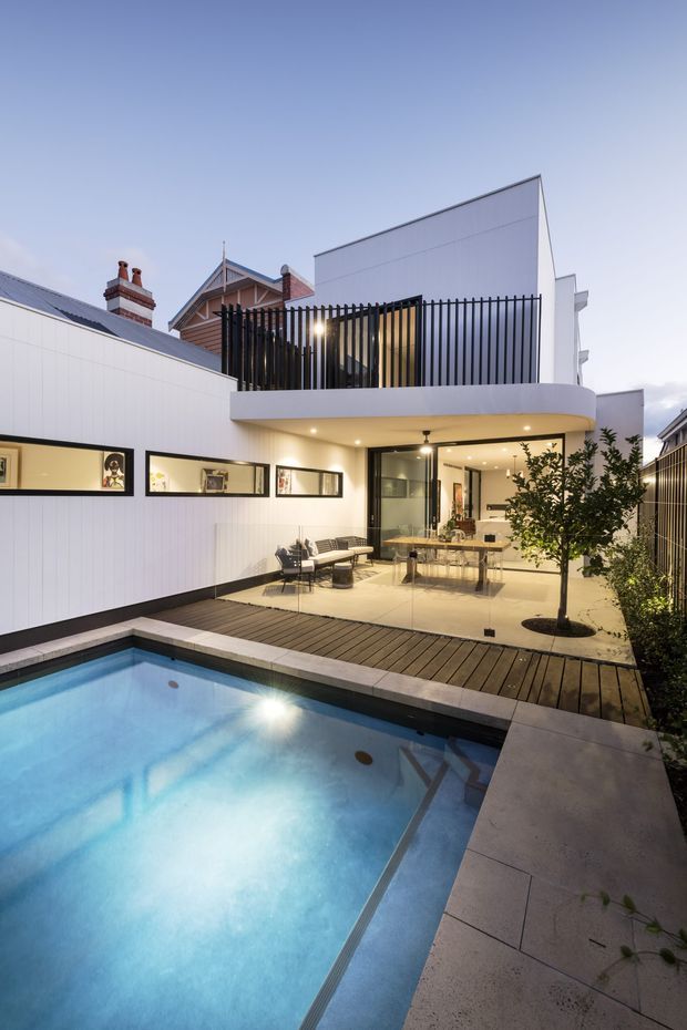 A contemporary multi-level residential extension viewed from a landscaped courtyard, with a rectangular swimming pool in the foreground, timber decking, pale tiled paving, and an illuminated glazed outdoor entertaining area beneath a projecting upper floor. The upper level features a black vertical-slat privacy screen and white cladding, while neighbouring pitched-roof homes are visible behind.