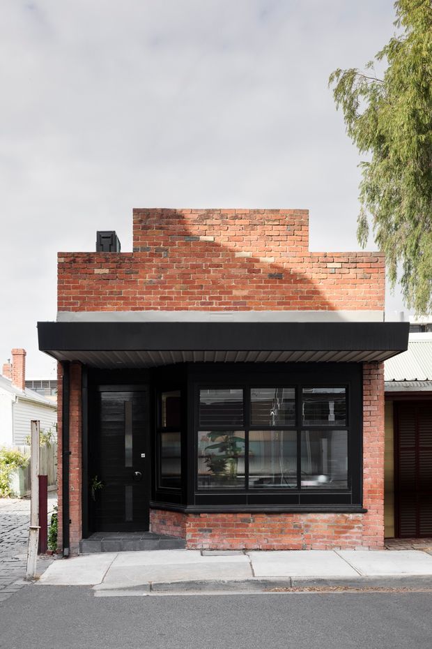 Street-facing view of a compact urban brick building with a flat black projecting canopy, dark entrance door, and broad black-framed window. The facade combines exposed red brickwork with black metal or painted frames and a simple contemporary composition. Adjacent low-rise buildings and a mature tree are visible at the edges.