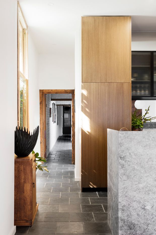 A bright interior hallway or entry passage with dark grey rectangular stone-like floor tiles, white walls, timber-framed glazing, and a prominent tall natural timber built-in cabinet. A large pale grey stone island or counter occupies the right foreground, with a black-framed glazed opening beyond. Warm timber trim and cabinetry contrast with the crisp white walls and dark flooring.