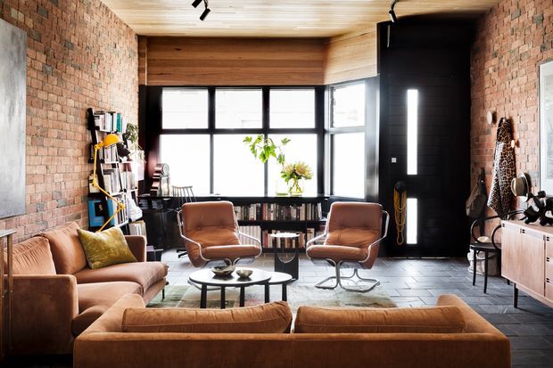 A warm, urban living room with exposed red brick walls, a timber-clad ceiling, black-framed floor-to-ceiling glazing, and a large black entry door. The room is furnished with tan leather lounge chairs and sofas, a round black coffee table, open shelving, a timber sideboard, and a compact dining area near the windows. Track lighting and a mix of timber, brick, and dark tiled surfaces create an industrial yet comfortable atmosphere.