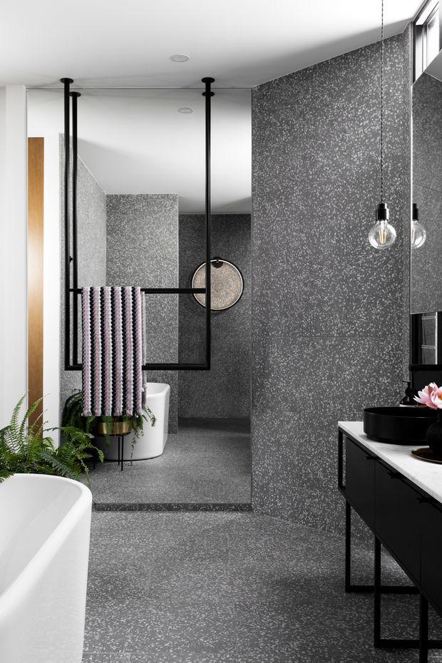 A contemporary bathroom with a freestanding white bathtub, dark speckled floor and wall tiles, a black floating vanity with a pale benchtop and round vessel basin, and a large wall mirror. Black-framed glass partitions, exposed pendant bulbs, a patterned towel, and indoor greenery add graphic accents to the monochrome space.