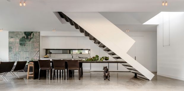 Bright open-plan interior featuring a dining area and contemporary kitchen beneath a dramatic sculptural white staircase. The space has polished light flooring, white built-in cabinetry, dark dining furniture, exposed dark stair treads, recessed lighting, and a large skylight or ceiling opening.