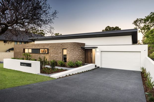 Contemporary single-storey suburban residence with a flat roof, white rendered walls, dark grey roof trim, warm exposed brick feature facade, timber entry door, and an integrated double garage. A broad asphalt driveway and landscaped front garden lead to the entry, with a low white boundary wall at the street.