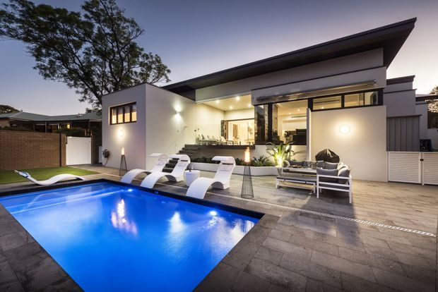 Contemporary single-storey home exterior at dusk, viewed from a landscaped pool terrace. The house features a sharply angled low-pitched roof, white rendered walls, extensive glazing, warm exterior lighting, and a timber-look entertaining deck. A brightly illuminated swimming pool, outdoor lounge furniture, sun loungers, and a planted courtyard create a polished resort-style setting.