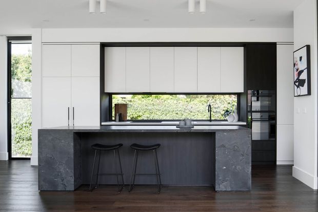 A refined contemporary kitchen with a large dark stone island, integrated seating for two, white full-height cabinetry, black-framed glazing, and a linear window overlooking greenery. Dark timber flooring and restrained black accents create a high-contrast, sophisticated interior.