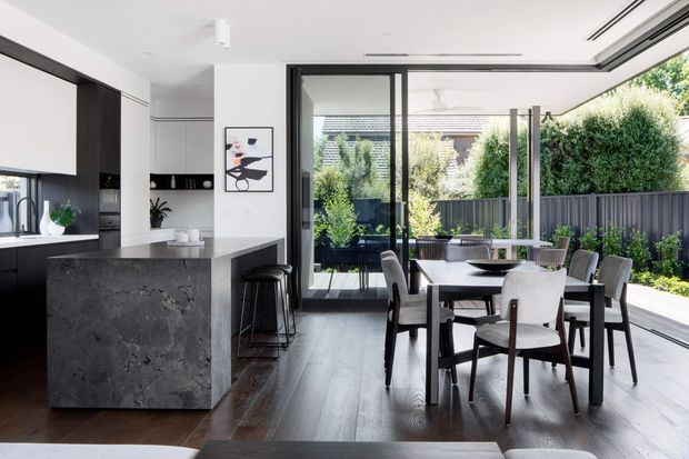 A bright, open-plan contemporary kitchen and dining area with a dark stone island, white cabinetry, timber flooring, and a rectangular dining table with upholstered chairs. Full-height black-framed glazing and sliding doors open toward a landscaped courtyard, bringing in abundant natural light.