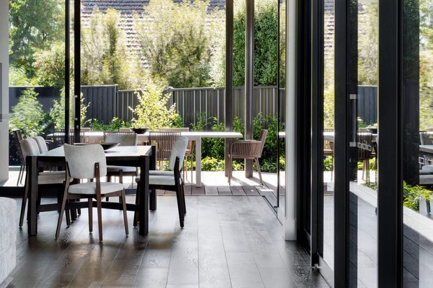 A bright contemporary dining area opening onto a landscaped outdoor terrace through extensive floor-to-ceiling glazing. The interior has a dark timber dining table with upholstered timber-framed chairs, pale large-format flooring, and black-framed glazed doors. Outside, a long narrow table, chairs, planter beds, and dark vertical fencing are visible, with greenery surrounding the space.
