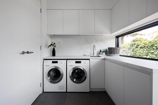 A bright, compact contemporary laundry room with two front-loading appliances beneath a white stone-look benchtop. Full-height pale grey cabinetry provides overhead storage and wraps around the room, while a deep white sink with a chrome mixer sits beside the appliances. A large horizontal window brings in natural light and overlooks greenery. The room has dark large-format floor tiles, clean handleless joinery, and a minimal monochromatic palette.