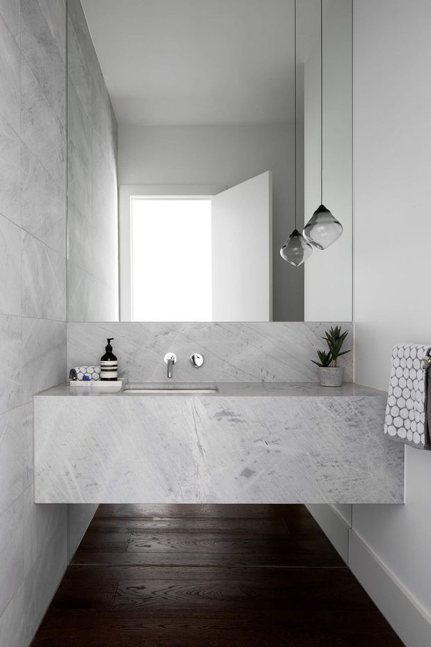 A narrow, refined contemporary bathroom featuring a wall-mounted stone vanity with integrated basin, large frameless mirror, pale veined stone wall and vanity surfaces, dark timber flooring, minimalist chrome fixtures, and sculptural clear pendant lights. A bright open doorway is visible in the mirror reflection.