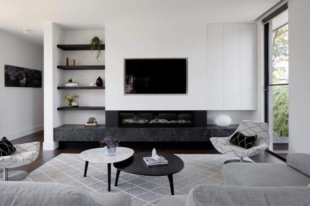 Contemporary living room with a monochrome palette, featuring a wall-mounted television, long dark stone fireplace surround and hearth, white built-in cabinetry, open display shelving, and large windows. A grey sectional sofa, patterned rug, black nesting coffee tables, and modern lounge chairs create a refined, comfortable seating area.
