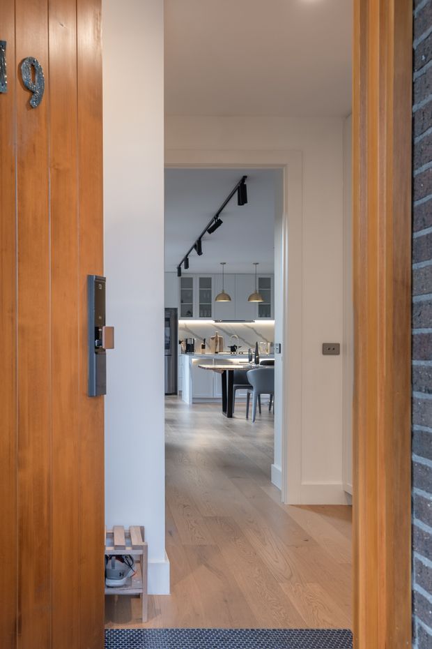 View from an open front entry into a bright contemporary home. A timber pivot or entry door and timber-framed opening sit beside exposed brickwork, while a pale timber floor leads through a hallway toward a kitchen and dining area. The kitchen features grey cabinetry, a light benchtop, pendant lights, and black track lighting. Shoes and a small entry bench are visible near the doorway.