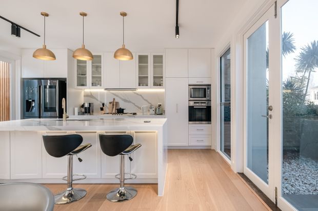 Bright contemporary kitchen with a white island and integrated seating for two, white shaker-style cabinetry, veined stone-look backsplash and benchtops, stainless-steel appliances, timber flooring, and three warm pendant lights. Full-height glazed doors and windows open toward a planted exterior area.