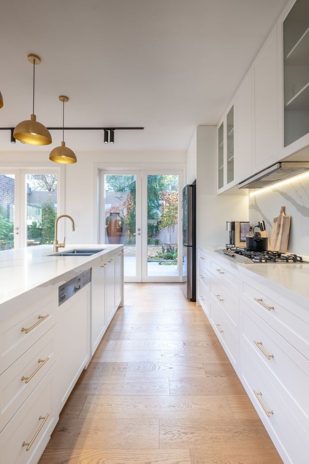 Bright, narrow galley-style kitchen with white Shaker cabinetry on both sides, a large white island with integrated sink and brass faucet, white stone benchtops, and a marble-look splashback. Light timber flooring, brass hardware, brass pendant lights, and black track lighting create a refined, contemporary transitional interior. Full-height glazed doors at the rear open to a garden courtyard, providing abundant natural light.
