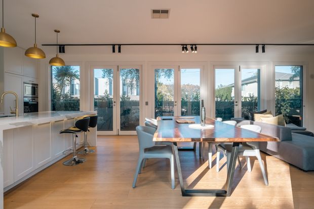 Bright open-plan kitchen and dining/living area with white cabinetry, a pale stone island and breakfast bar, timber flooring, a timber dining table, and contemporary pendant and track lighting. Full-height glazed doors open to an outdoor area, creating an airy connection to the garden.