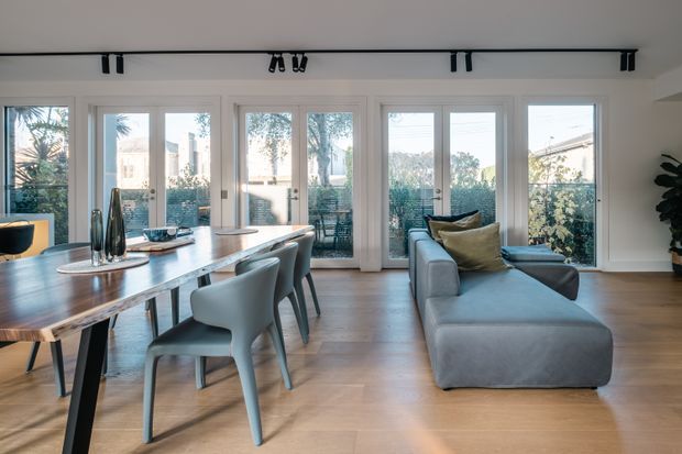 Bright, open-plan contemporary living and dining area with a long timber dining table and upholstered dining chairs in the foreground, and a large grey sectional sofa facing away from the camera. Full-height multi-panel glazed doors open to an outdoor terrace or garden, bringing in abundant natural light. Track lighting runs across the ceiling, with simple white walls, warm timber flooring, and restrained neutral furnishings.