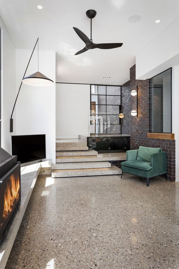 A bright, contemporary open-plan living area with polished terrazzo flooring, a split-level stepped floor, and a dramatic exposed dark brick feature wall. The room includes a black-framed glazed partition, a wall-mounted television, a linear fireplace, sculptural pendant lighting, a black ceiling fan, and a green upholstered lounge chair. White walls and ceiling create a clean, airy contrast with the darker brick and joinery.