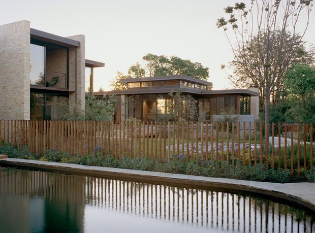 A contemporary low-rise residential house viewed from across a landscaped water feature. The home has pale brick masonry walls, extensive dark-framed glazing, strong horizontal roof planes and a timber slat boundary fence, set among mature trees and planting.