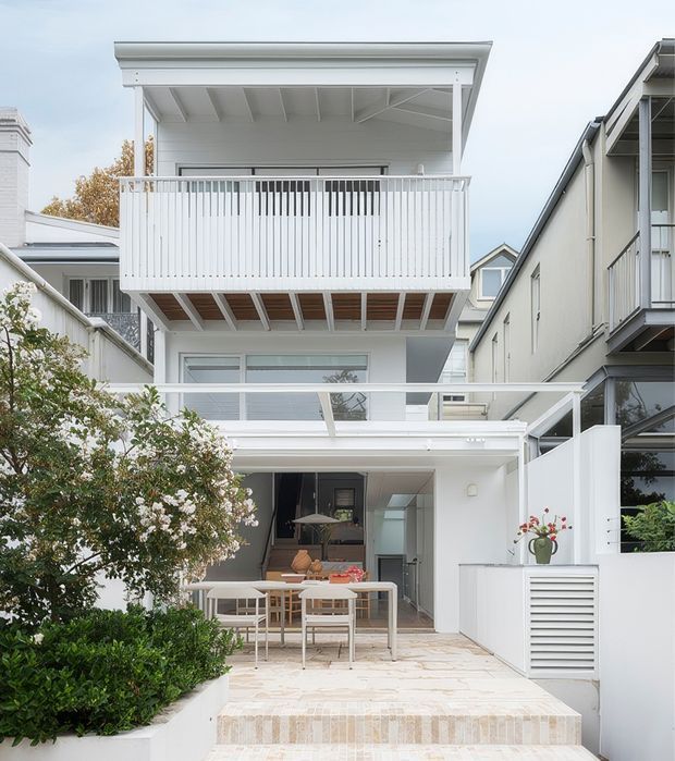 A bright white multi-storey residential facade viewed from a landscaped outdoor patio. The home features layered balconies with slim vertical white balustrades, exposed timber-like balcony soffit framing, a covered ground-floor entertaining area with outdoor dining furniture, pale stone or tiled paving and steps, and clean modern coastal detailing. Neighbouring attached residences are visible on either side.