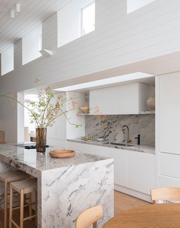 Bright, refined contemporary kitchen with white panelled walls and ceiling, tall clerestory-style windows, extensive white cabinetry, and a large waterfall island clad in dramatic veined natural stone. A matching stone splashback surrounds the sink, with warm timber flooring and timber bar and dining seating adding contrast. The open-plan space has an airy, high-ceilinged feel and includes a dining area in the foreground.
