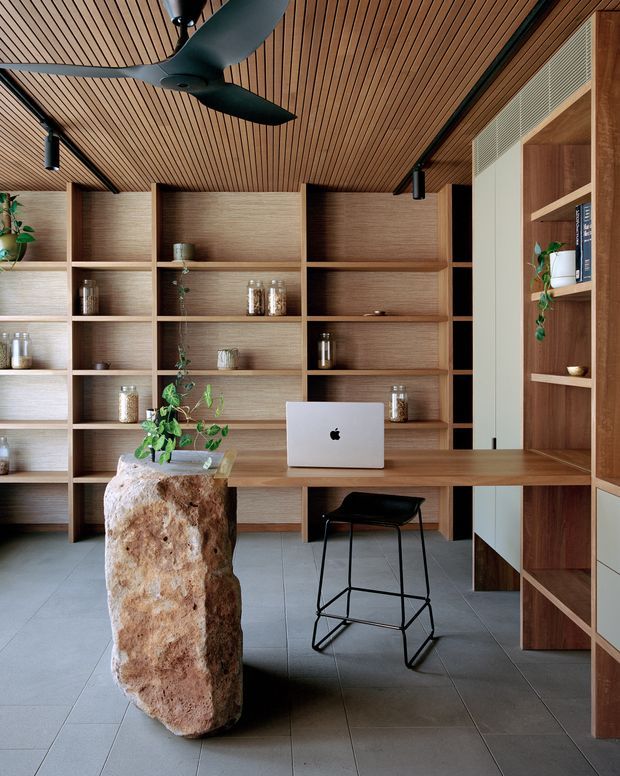 A refined home office with extensive built-in timber shelving and a long timber desk, furnished with a laptop and a simple black stool. The room features a slatted timber ceiling, exposed black track-style lighting, grey tiled flooring, integrated white cabinetry, and a large rough natural stone pedestal supporting a plant. The overall design is warm, contemporary, and highly crafted, with strong custom joinery and biophilic accents.
