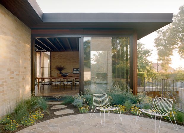 Contemporary residential patio courtyard viewed from outside, with pale stone paving, native-style planting, white wire-frame outdoor chairs, and expansive dark-framed glazing opening to a dining area. The low-slung flat roof, exposed timber soffit, and warm masonry walls create a refined indoor-outdoor connection.