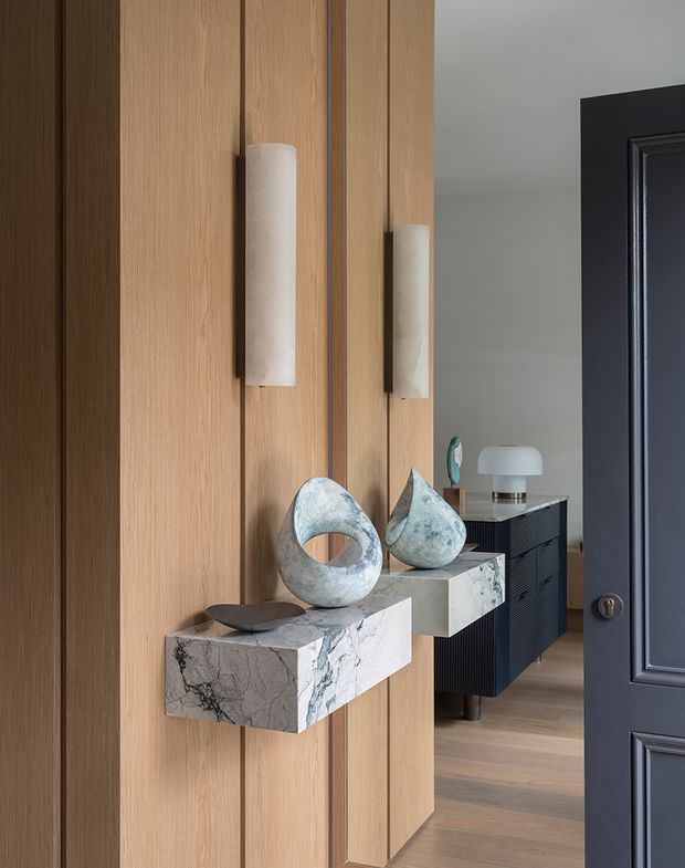 A refined interior entryway featuring a full-height fluted timber wall with two sculptural wall sconces, floating veined stone display ledges, and decorative ceramic or stone objects. A dark paneled door opens toward a timber-floored adjoining space containing a dark console cabinet and additional decor. The composition is warm, sophisticated, and carefully curated.
