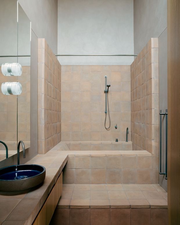 A warm, minimalist bathroom featuring a large tiled walk-in shower with partial-height tiled privacy walls, a built-in tiled bath or ledge, a dark vessel basin on a timber-toned vanity, exposed dark metal tapware, and a full-height mirror. The space uses earthy beige and terracotta tilework with restrained contemporary detailing.