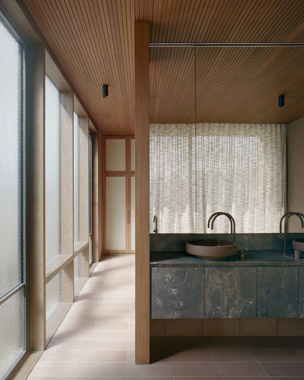 A refined bathroom vanity area viewed beside a glazed corridor. The space features a dark stone vanity with integrated cabinetry, vessel basins and curved black faucets, backed by textured translucent blinds. Warm timber slat ceilings, timber framing, frosted floor-to-ceiling windows and pale tiled flooring create a calm, softly lit interior.