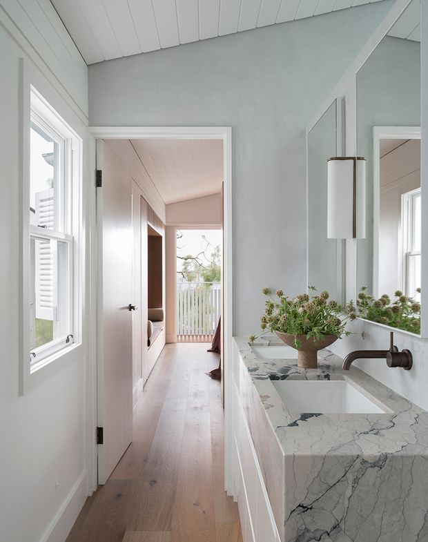 A bright, refined bathroom viewed from the vanity area, featuring a white marble vanity top with an integrated rectangular basin, dark bronze wall-mounted faucet, large mirror, and cylindrical wall sconce. The room has pale blue-grey painted walls, white vertical wall panelling and trim, a light timber floor, and a doorway opening to a light-filled hallway or adjoining bedroom area.