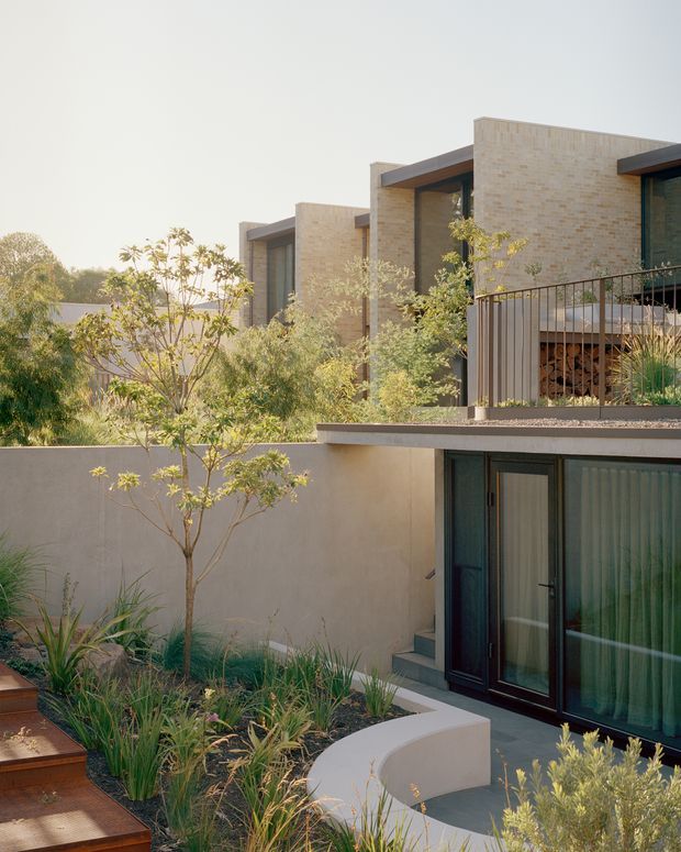 Exterior view of a contemporary multi-level residence with warm light brick walls, deep roof overhangs, dark-framed glazing, and layered terraces. A landscaped courtyard garden with ornamental grasses, small trees, curved rendered walls, and timber steps occupies the foreground.