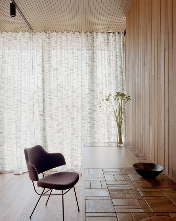 A refined, warm-toned interior corner featuring a sculptural upholstered lounge chair beside a minimalist built-in desk or console. Full-height sheer curtains diffuse daylight across a light timber floor, while vertical timber wall panelling and a matching slatted ceiling create a cohesive natural-material envelope. The desk surface displays a simple vase of dried stems and a dark bowl, with a patterned rug grounding the seating area.