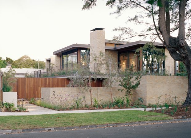 Street-facing view of a contemporary two-level residence with low-pitched rooflines, extensive glazing, pale stone masonry walls, timber screening, a prominent rectangular chimney, and landscaped garden edges. Mature trees partially frame the facade.